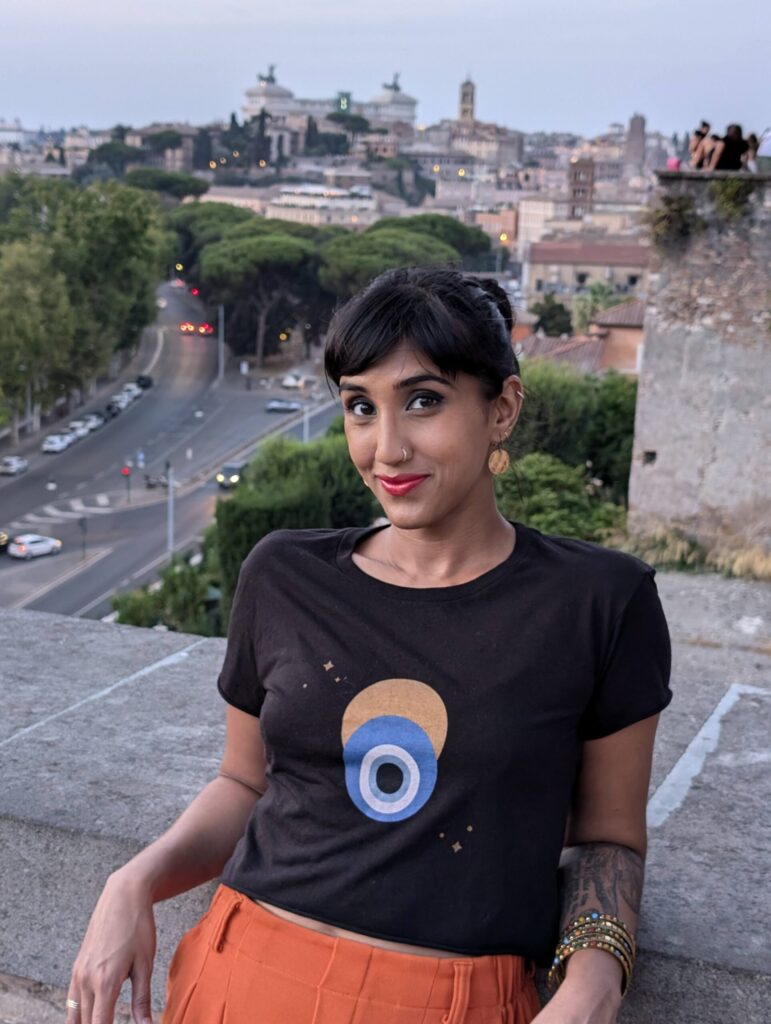 A woman with dark hair, red lipstick, and a printed t-shirt poses casually on a stone wall overlooking a cityscape with historic buildings, trees, and winding roads at dusk.
