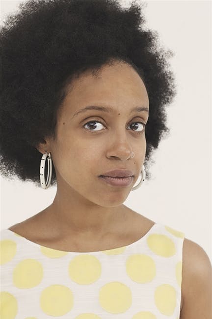 A woman with natural curly hair wears large hoop earrings and a white top with yellow polka dots, looking calmly at the camera against a plain background.