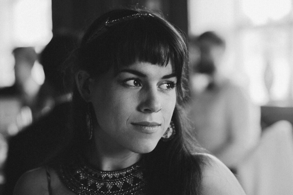 A black-and-white portrait of a woman with long dark hair and bangs, wearing a beaded necklace and earrings, looking thoughtfully to the side. The background is softly blurred with indistinct figures.
