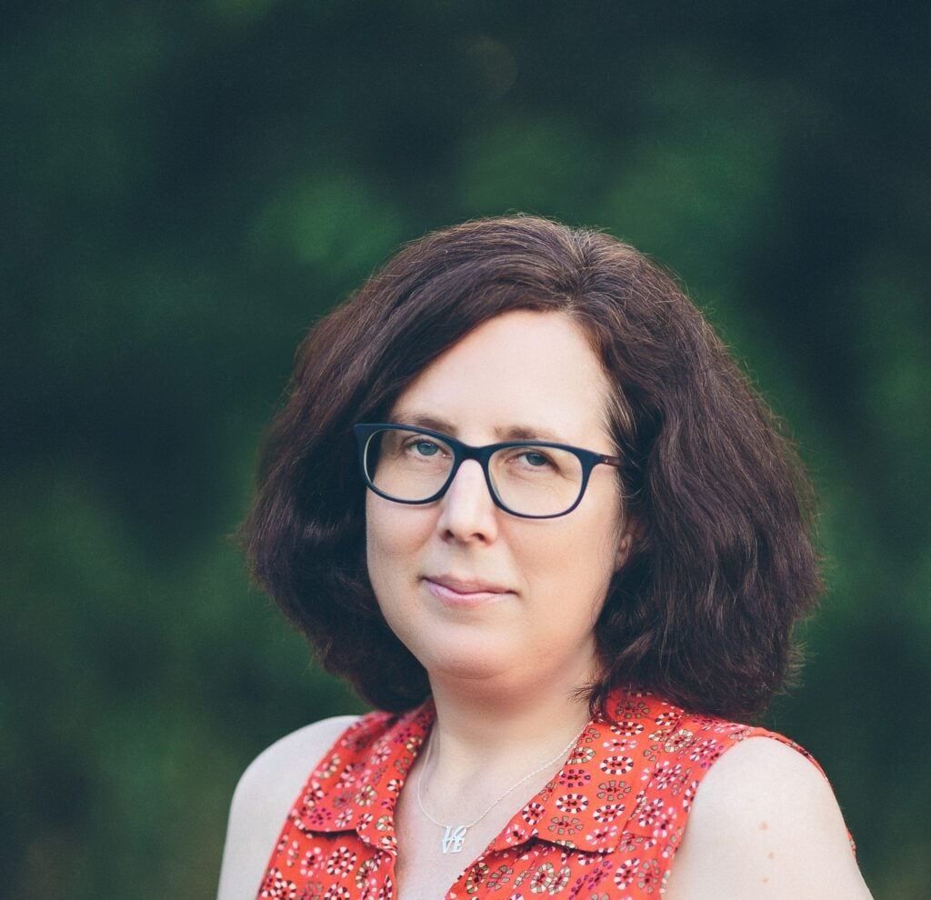 A woman with shoulder-length brown hair and glasses wears a red patterned sleeveless top, standing outdoors with a blurred green background. She looks at the camera with a neutral expression.