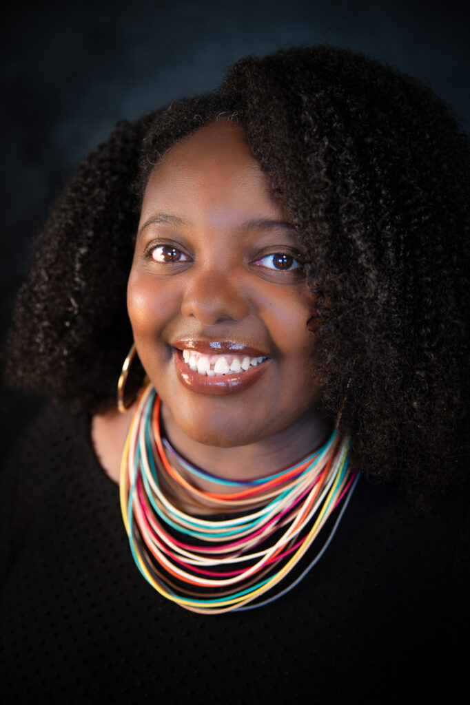 A smiling woman with curly hair wears a black top, large hoop earrings, and a colorful multi-strand necklace, posing against a dark background.