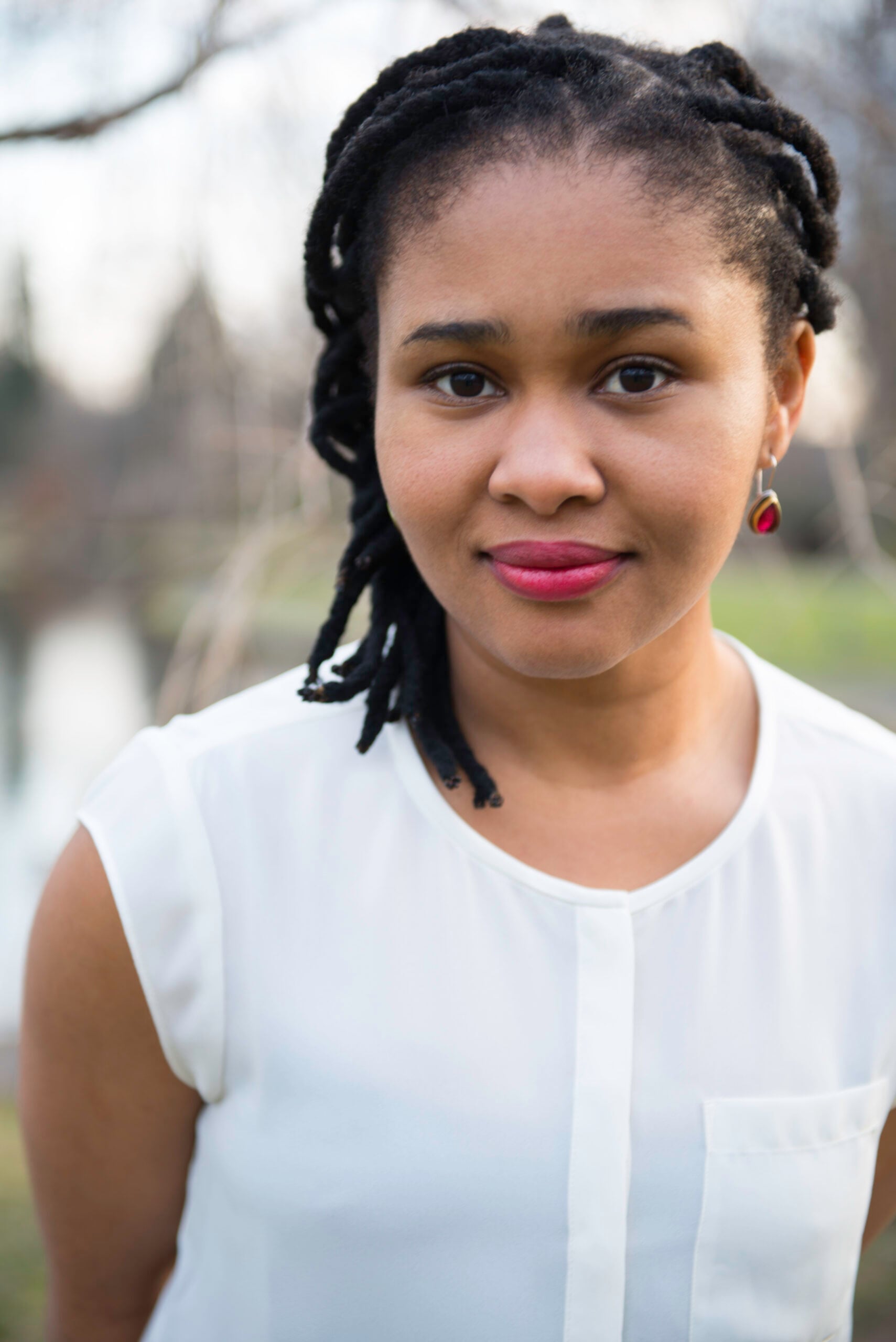 A woman with black dreadlocks, wearing a white sleeveless blouse and red earrings, stands outdoors with a soft smile. The background is blurred, showing greenery and water.