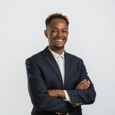 A smiling man wearing a dark suit jacket and white shirt stands with arms crossed against a plain white background.