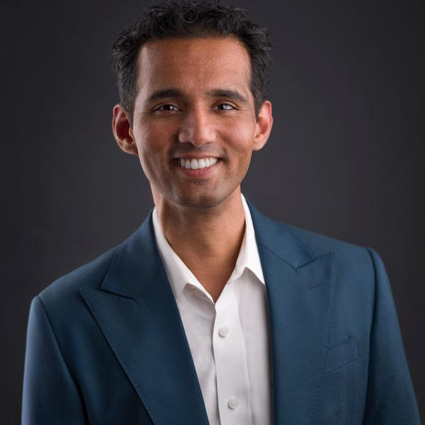 A man with short, dark hair smiles at the camera while wearing a white shirt and a blue suit jacket against a dark background.