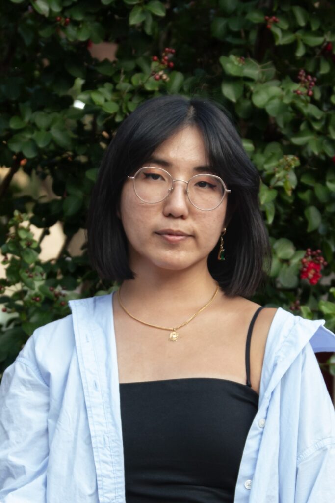 A young person with straight black hair, round glasses, and a neutral expression wears a black top, light blue shirt, and gold necklace, standing in front of a leafy green background with red berries.
