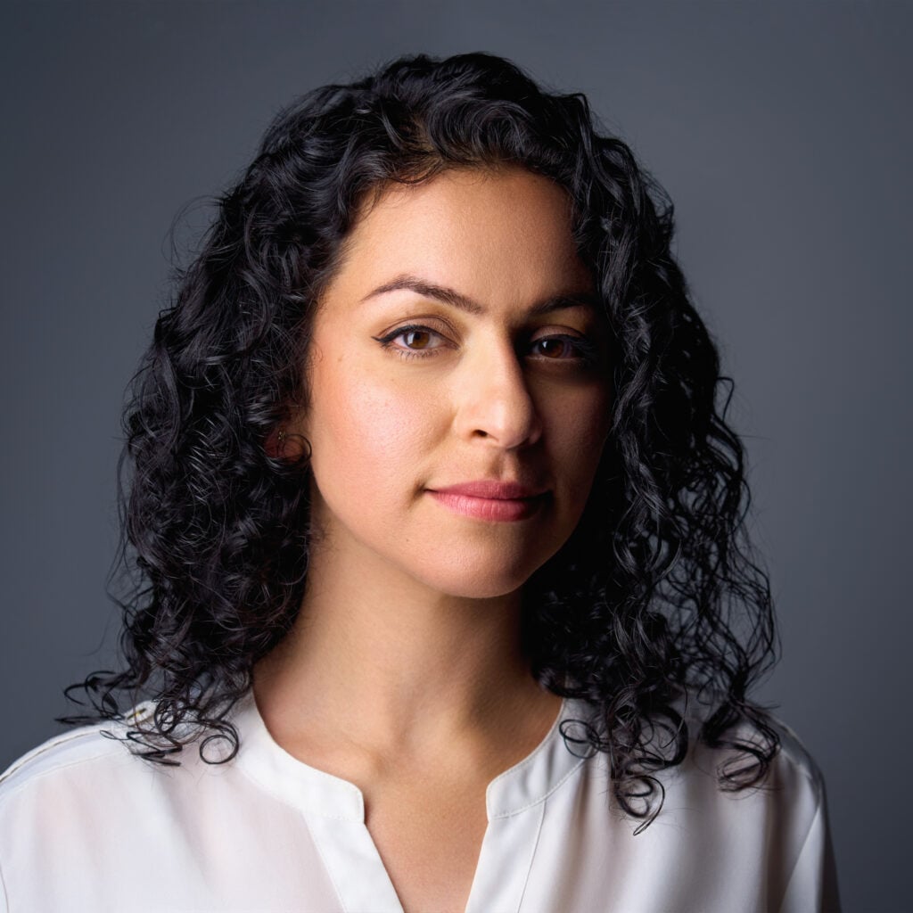 A woman with curly dark hair and medium skin tone, wearing a white blouse, looks confidently at the camera against a plain gray background.