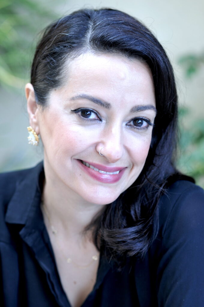 A woman with long dark hair, wearing a black top, gold earrings, and a nose ring, smiles warmly at the camera with a blurred green background.