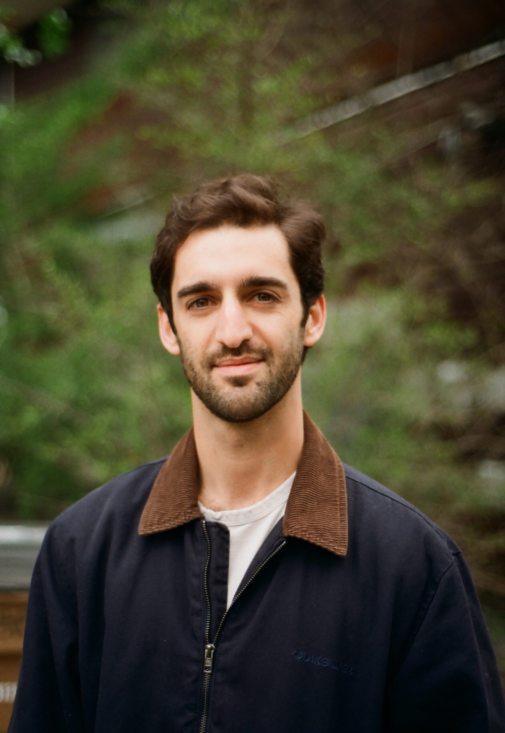A man with dark hair and a trimmed beard stands outdoors, wearing a navy jacket with a brown collar and a white shirt. Green foliage and a blurred background are visible behind him.