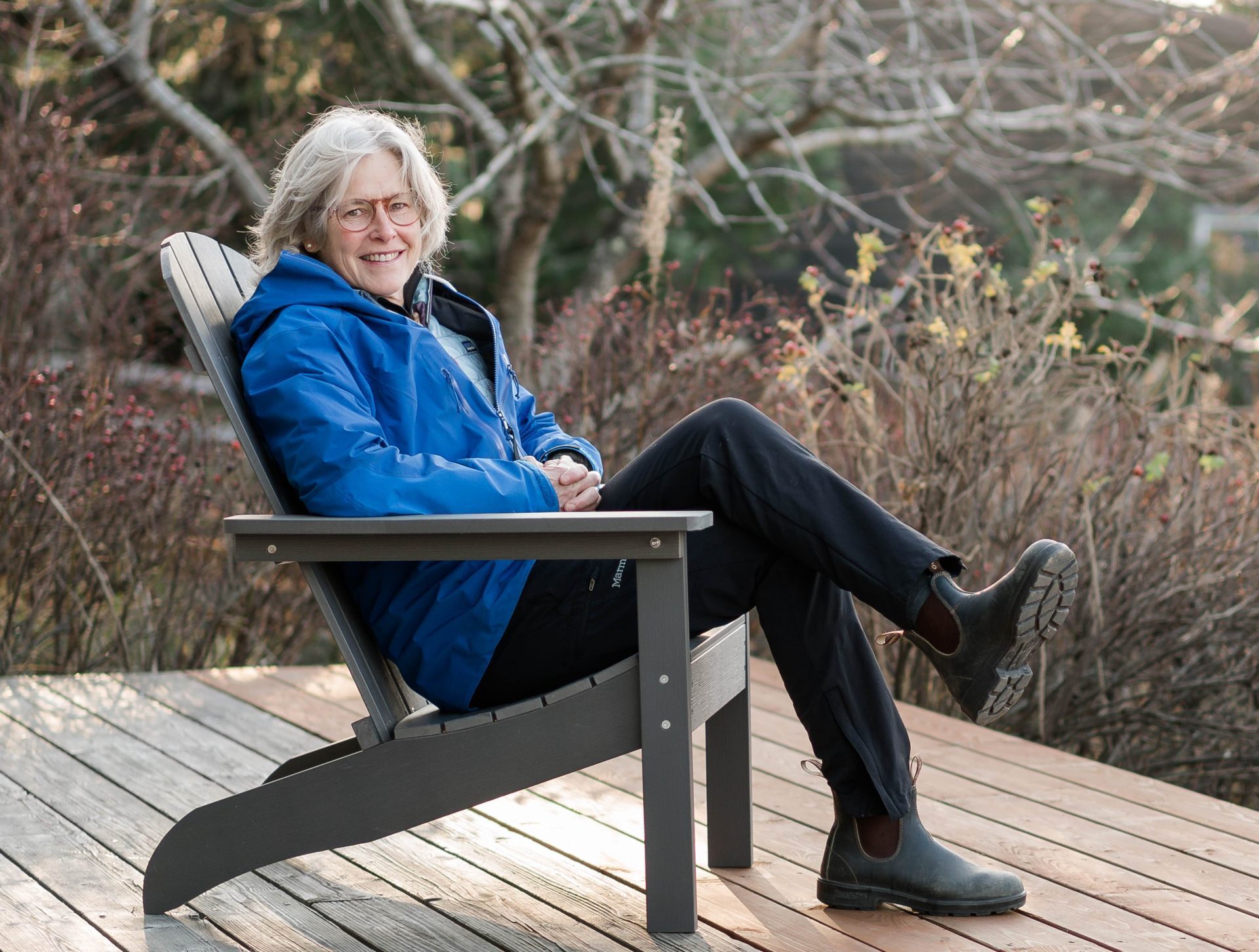 An older woman with gray hair and glasses, wearing a blue jacket, dark pants, and boots, sits smiling in a wooden chair on a deck outdoors, surrounded by bare trees and bushes.