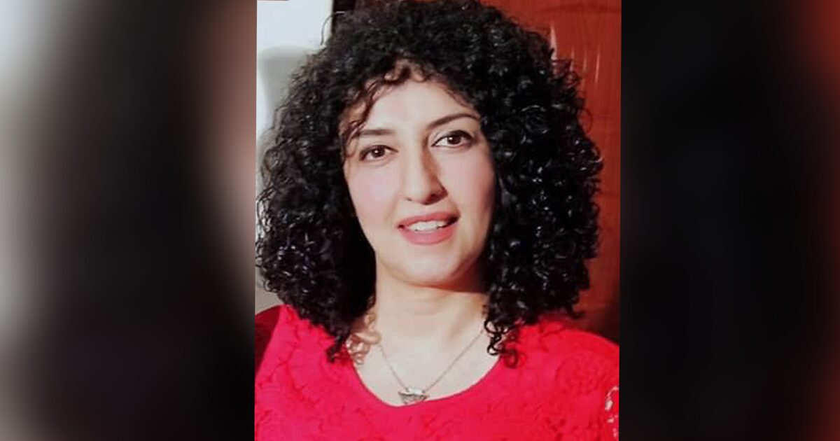 A woman with curly dark hair, wearing a bright red top and a necklace, smiles softly at the camera against a blurred indoor background.