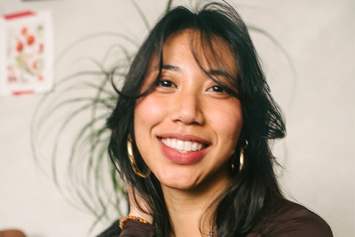 A woman with long dark hair and gold hoop earrings smiles warmly at the camera. She is indoors, with a blurred background featuring a leafy plant and a colorful picture on the wall.