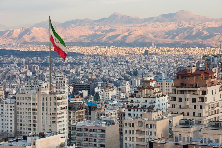 A large Iranian flag flies above tall buildings in Tehran, with a sprawling cityscape and distant mountains visible under a soft, early evening light.