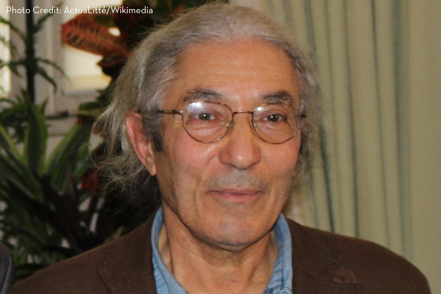 An older man with long gray hair, glasses, and a brown jacket stands indoors in front of a plant and beige curtains. Photo credit: ActuaLitté/Wikimedia.