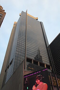 A tall glass skyscraper with a pointed rooftop stands against a cloudy sky. At the base, a digital billboard displays a person wearing headphones, speaking into a microphone.