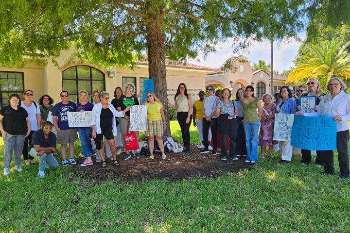 A diverse group of adults stands outside a building, holding signs with messages like Let Us Read and Let Our Kids Read. They appear to be peacefully protesting or advocating for reading and education.