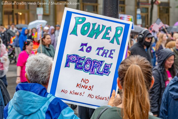 A person at a crowded outdoor protest holds a colorful sign reading “Power to the People. No Kings! No Kings! No Kings!” People in jackets and rain gear are visible in the background.