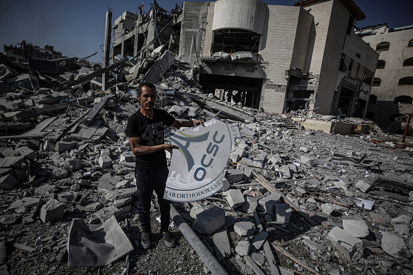 A man stands amid rubble, holding a damaged sign with OCSC text. Behind him, extensive destruction is visible, with collapsed buildings and debris scattered everywhere under a cloudy sky.
