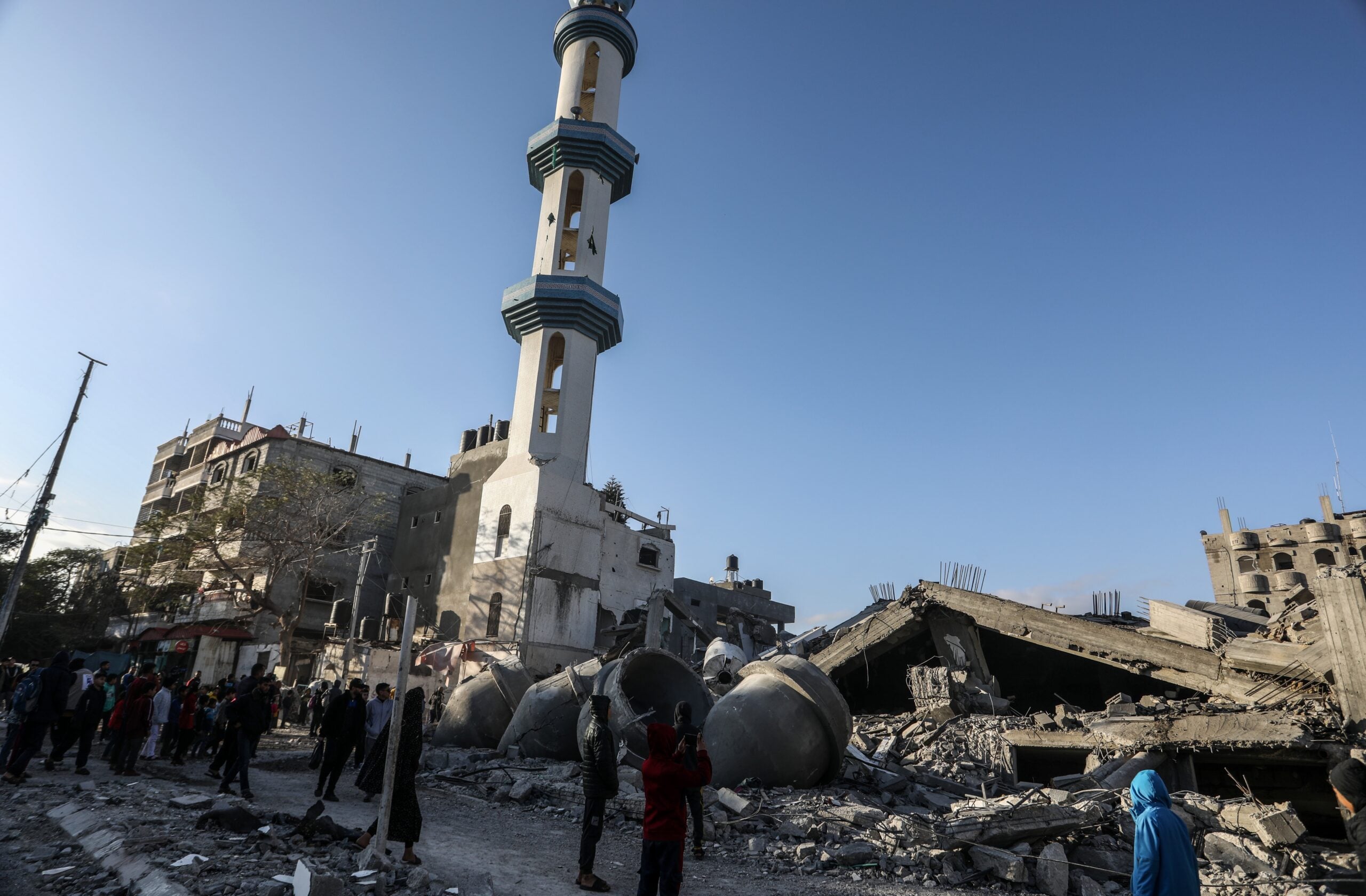 A group of people stand near the rubble of a destroyed building beside a tall mosque minaret under a clear blue sky. Debris and large concrete pieces are scattered across the ground.