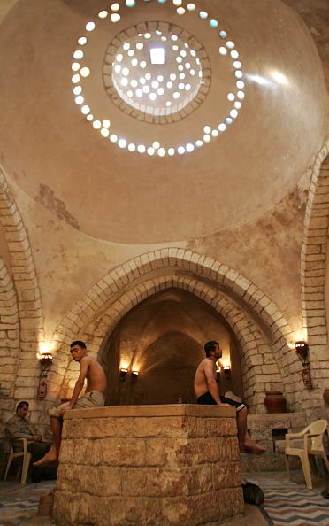Shirtless men sit on a stone platform under a large domed ceiling with circular skylights, inside a historic, arched bathhouse with beige stone walls.