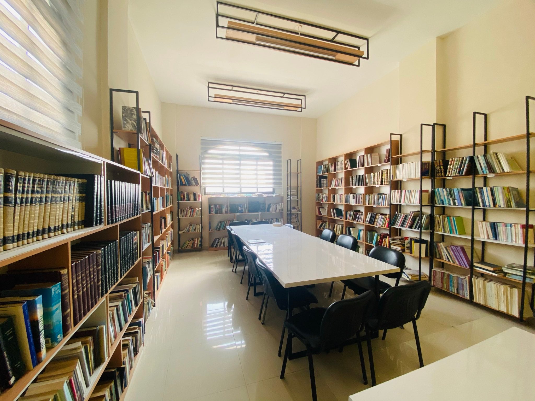 A bright library room with shelves full of books on both sides, a large white table in the center surrounded by black chairs, and sunlight streaming in through a window at the far end.
