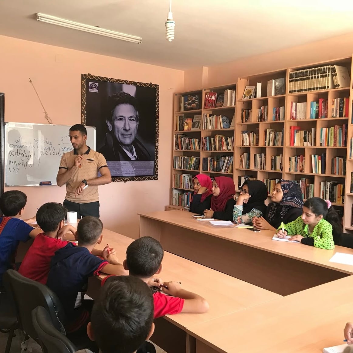 A teacher stands by a whiteboard teaching a group of children seated at tables in a library. The children are listening attentively. Bookshelves line the walls and a large portrait hangs in the background.