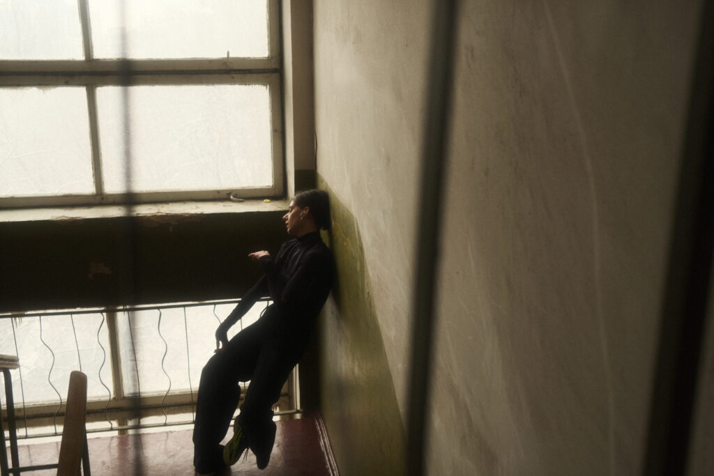 A person in dark clothing leans against a wall in a dimly lit room, next to a large window with soft light coming through, creating a moody and contemplative atmosphere.