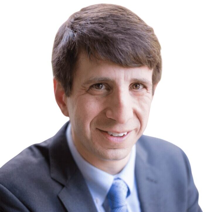 A man in a suit and blue tie smiles at the camera with his arms crossed. He has short brown hair and is posed against a plain white background.