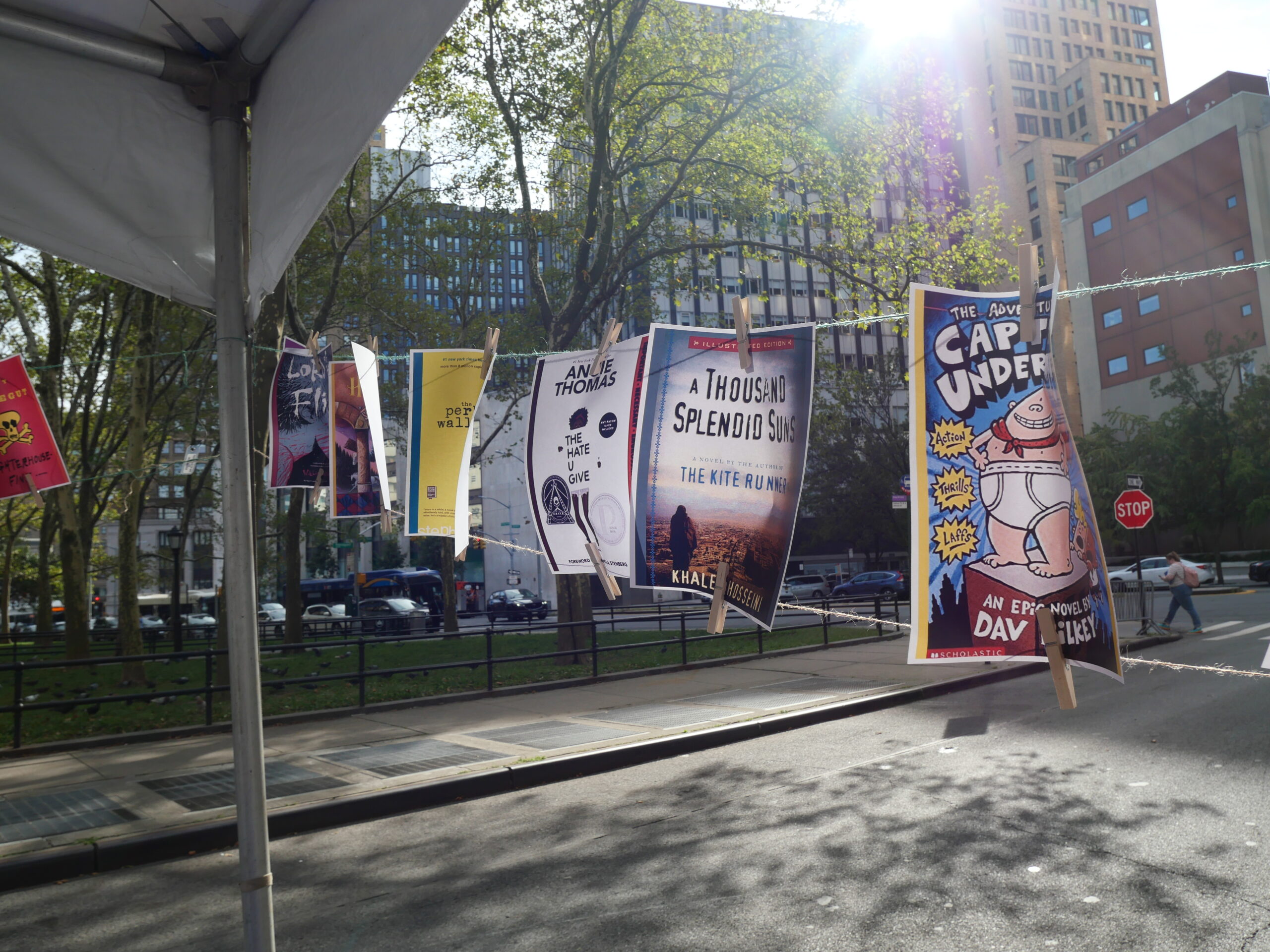 Colorful book covers hang on a line outdoors in a city park, with sunlight streaming through trees and tall buildings in the background. A street and stop sign are visible nearby.