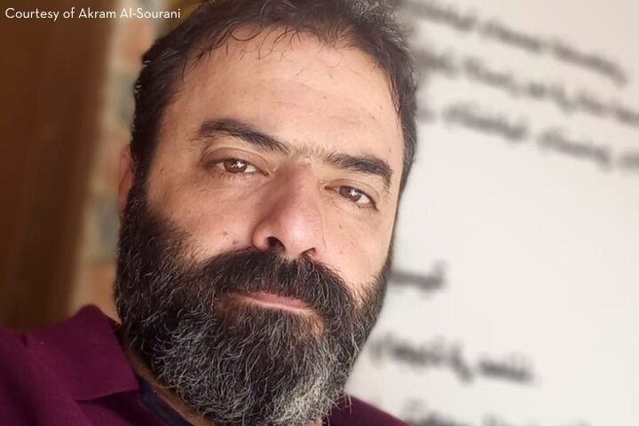 A man with a beard and mustache looks at the camera, standing indoors in front of a white wall with partially blurred Arabic writing. Courtesy of Akram Al-Sourani appears in the top left corner.