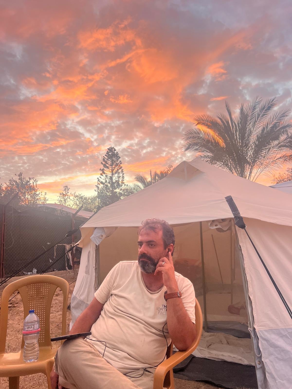 A man sits pensively on a plastic chair in front of a white tent at sunset, with vibrant orange clouds in the sky, palm trees, and a water bottle on a small table beside him.