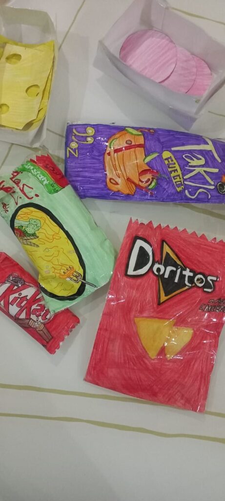 Hand-drawn paper replicas of snack packages, including Doritos, KitKat, Takis, a green chip bag, and a yellow cheese wedge, are displayed on a white surface with lines.