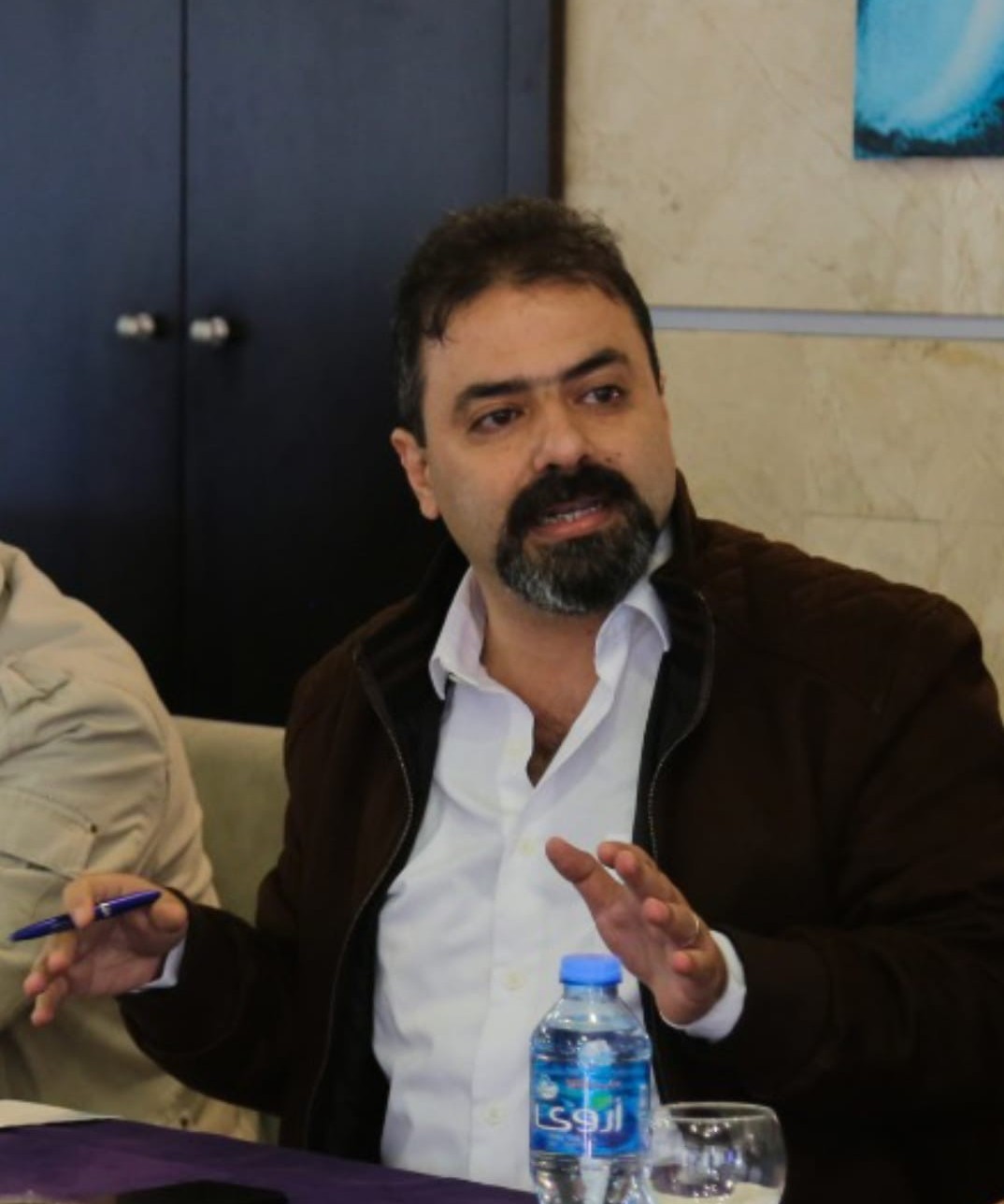 A man with dark hair and a beard, wearing a white shirt and brown jacket, gestures with one hand while holding a pen. He is seated at a table with a water bottle and glass in front of him.