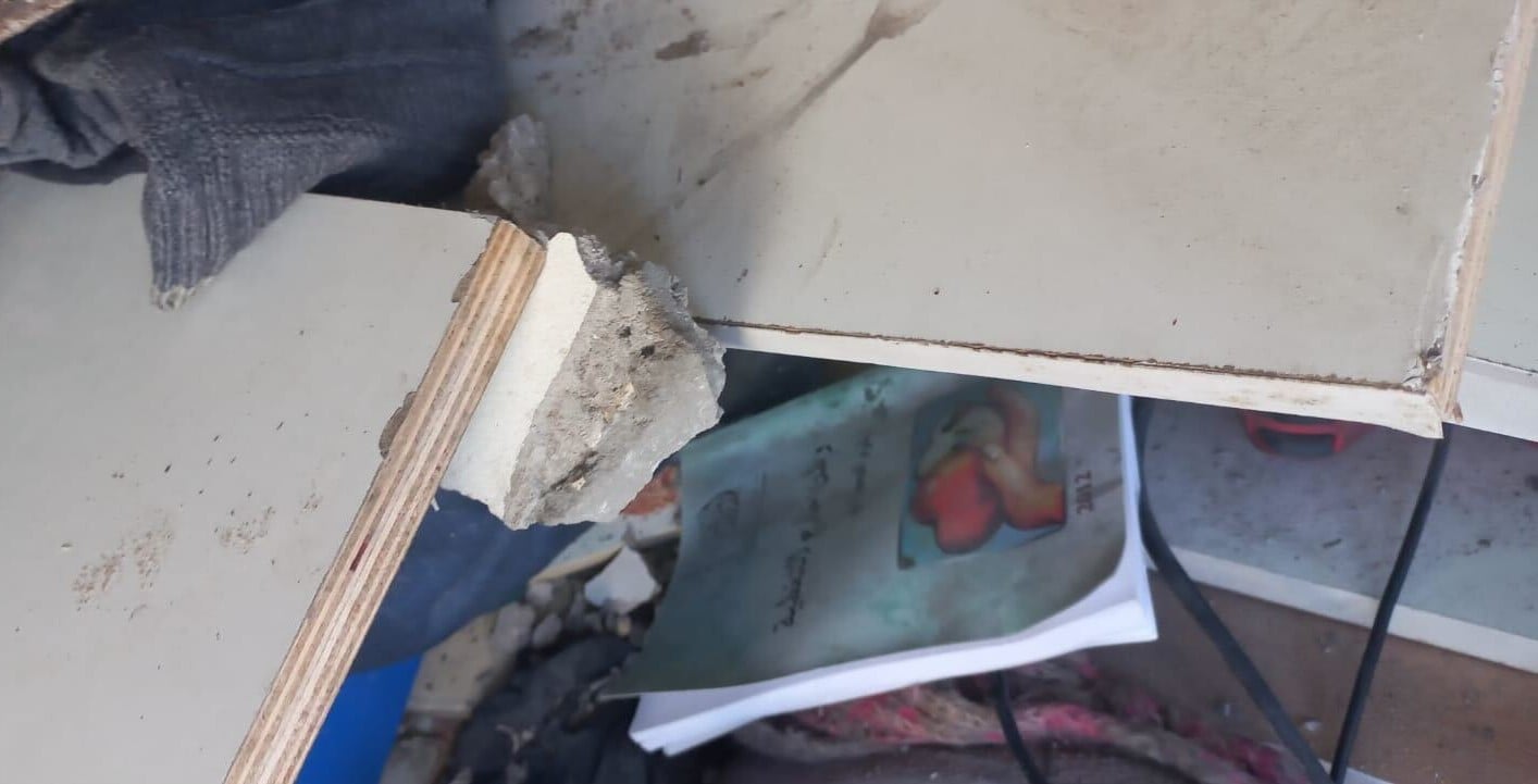 A broken wooden shelf with rough edges lies on a cluttered surface. Below it, a book with an illustrated cover and Arabic text is partially visible among scattered debris and other items.