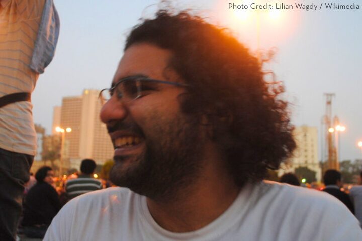 A man with curly hair and glasses smiles while sitting outdoors in a city at dusk, with blurred buildings and people in the background.