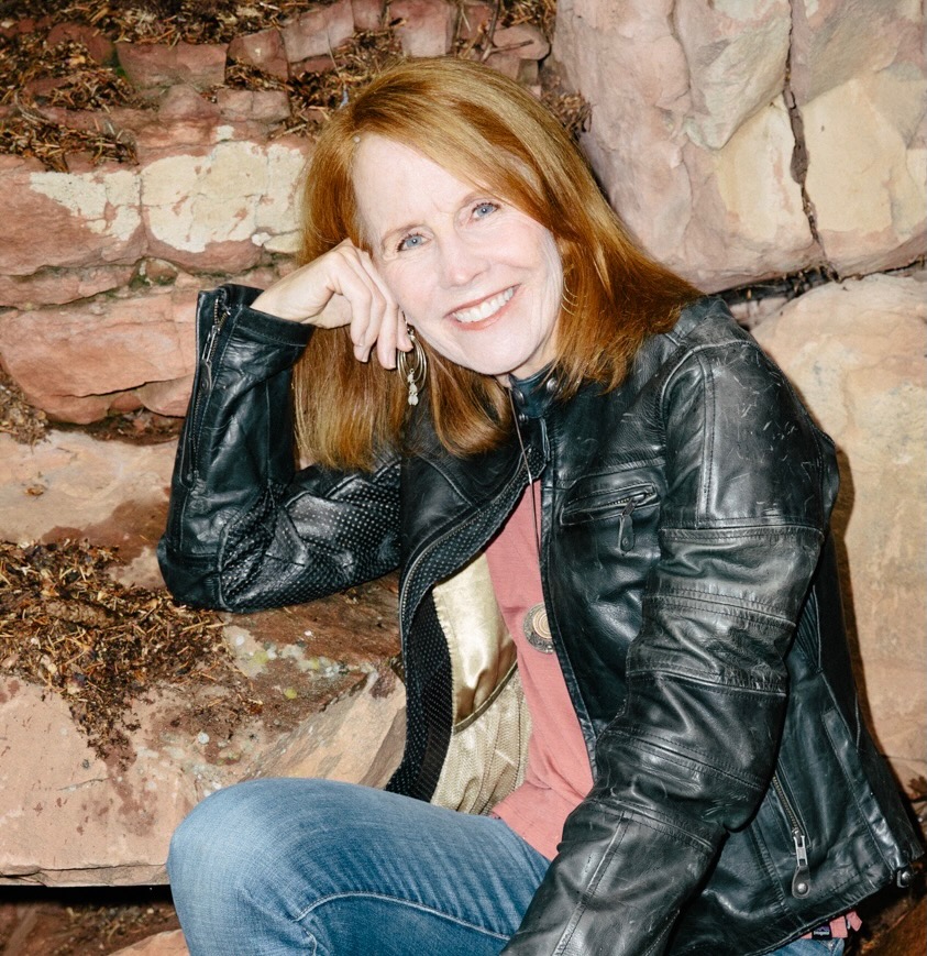 A woman with red hair wearing a black leather jacket and blue jeans sits on stone steps, smiling and resting her head on her hand, with a rocky background behind her.