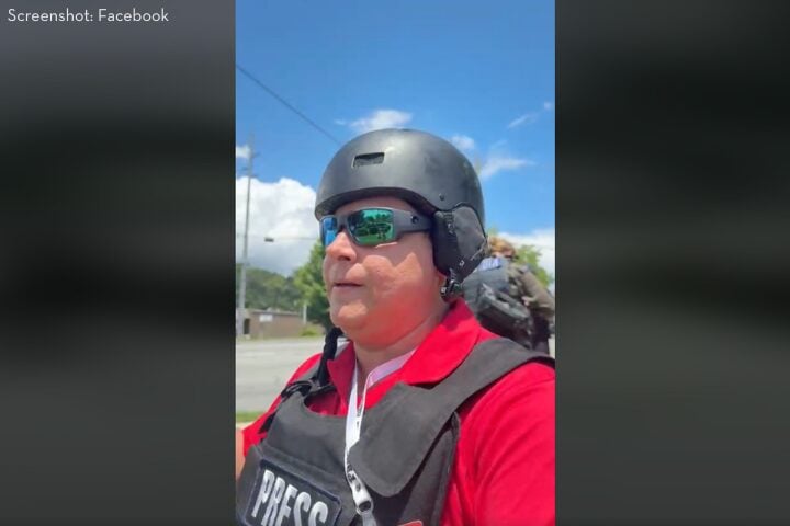 A person wearing a black helmet, sunglasses, and a vest labeled PRESS stands outdoors on a sunny day, with blue sky and clouds visible in the background.