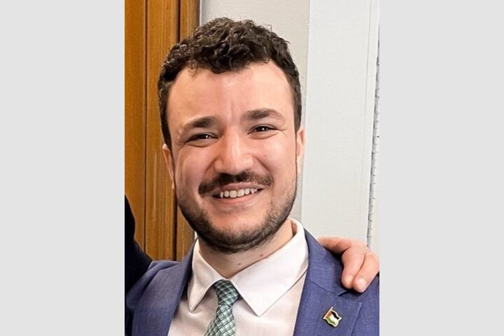 A man with short curly hair and a beard smiles at the camera, wearing a blue suit, white shirt, and light green tie. A small flag pin is on his lapel, and an arm rests on his shoulder.