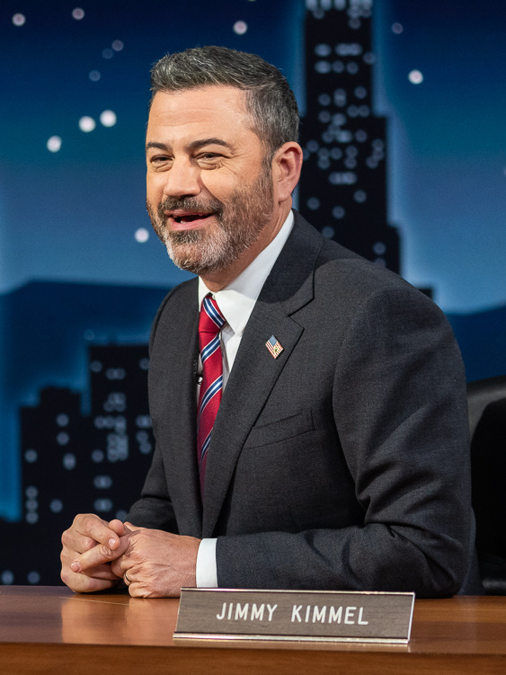 A man in a suit and striped tie sits at a desk with a nameplate reading Jimmy Kimmel, smiling and talking. The background shows a night cityscape.