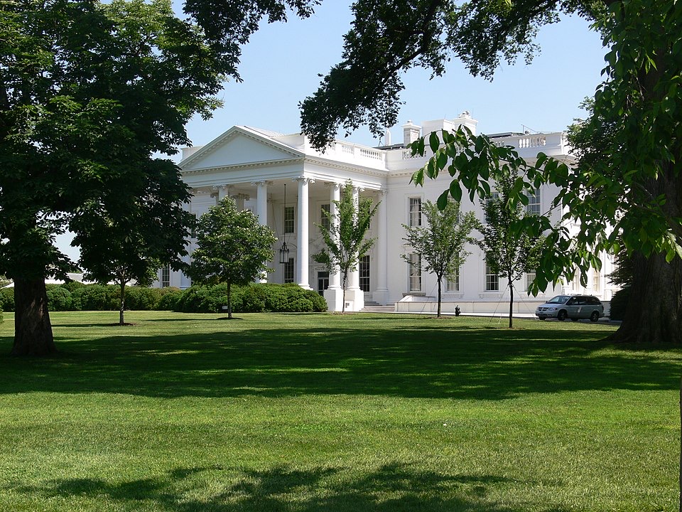 The White House is seen through leafy trees, with its white columns and portico visible. A car is parked nearby on the lush green lawn under a clear blue sky.
