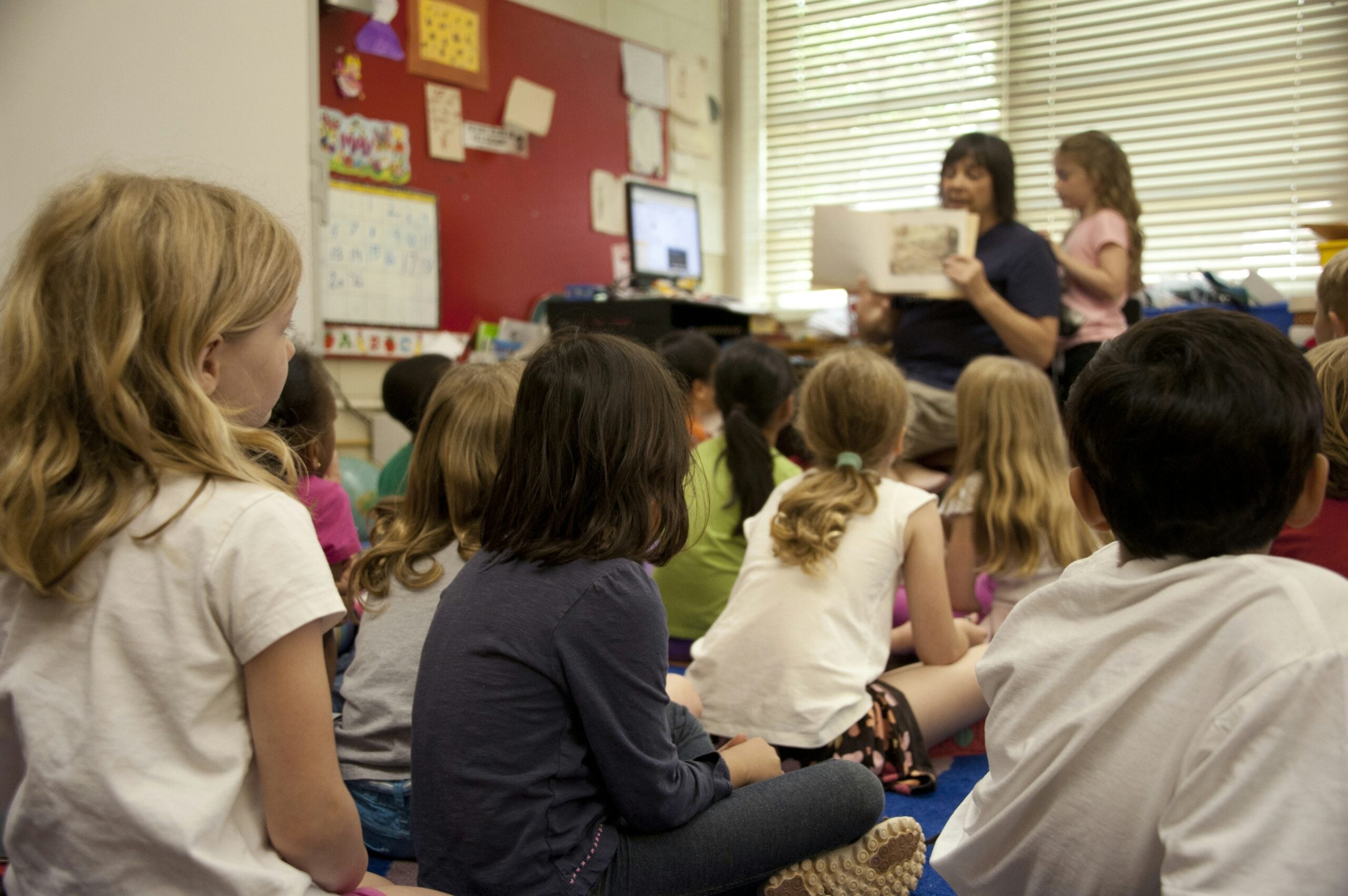 A group of young children sit on the floor in a classroom, attentively listening to a teacher at the front who is reading a book aloud. Sunlight streams through the window blinds behind the teacher.