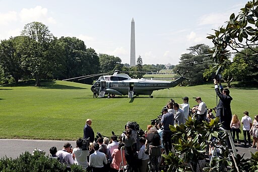 A helicopter labeled “United States of America” is parked on the White House lawn as photographers and people gather nearby, with the Washington Monument visible in the background.