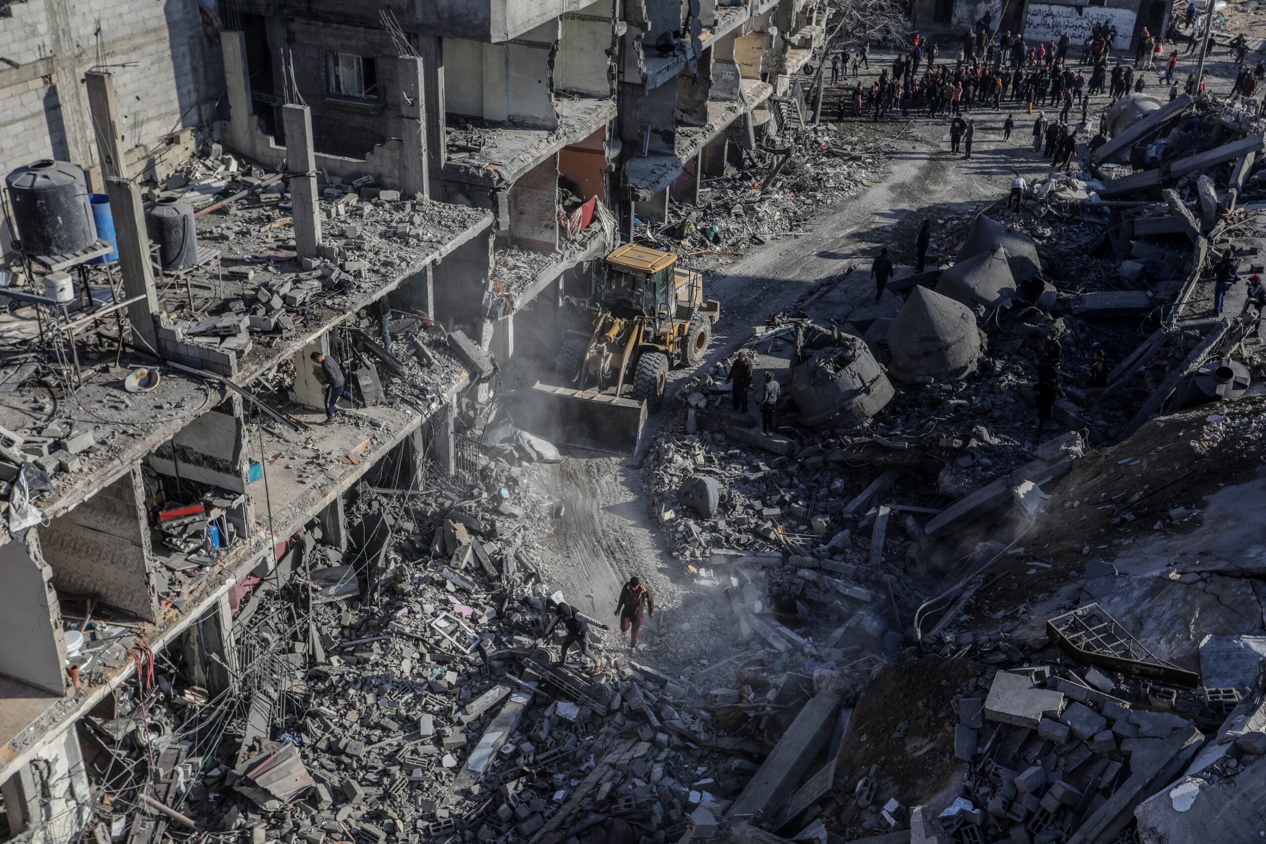 A bulldozer clears rubble from heavily damaged buildings and debris-strewn streets, while people survey the destruction and gather in the background.