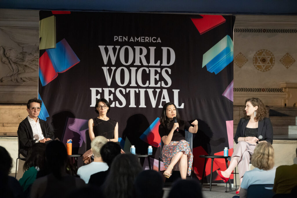 Four panelists sit on stage in front of a PEN America World Voices Festival banner, engaging with an audience. One woman speaks into a microphone while the others listen attentively.