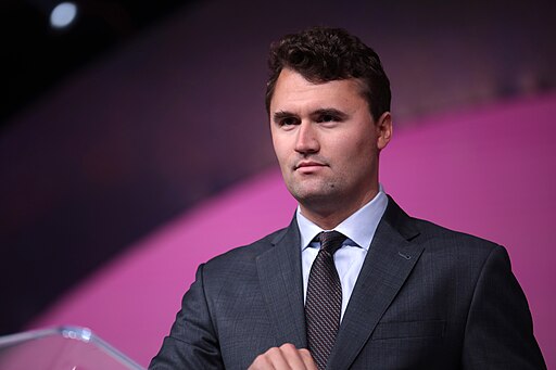 A man in a dark suit and tie stands in front of a purple and pink background, looking forward with a neutral expression.