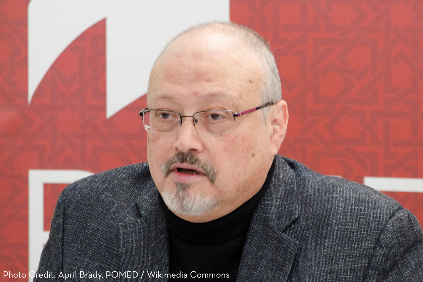 A middle-aged man with glasses, a goatee, and a bald head wears a dark blazer and black shirt, speaking in front of a red patterned background. Photo credit: April Brady, POMED / Wikimedia Commons.