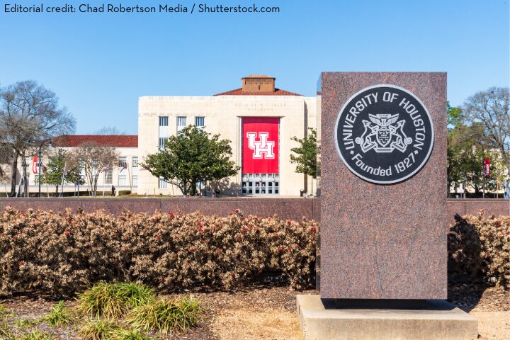 A large University of Houston stone monument stands in front of a campus building with a red UH banner. Trees, shrubs, and clear blue skies are visible in the background.