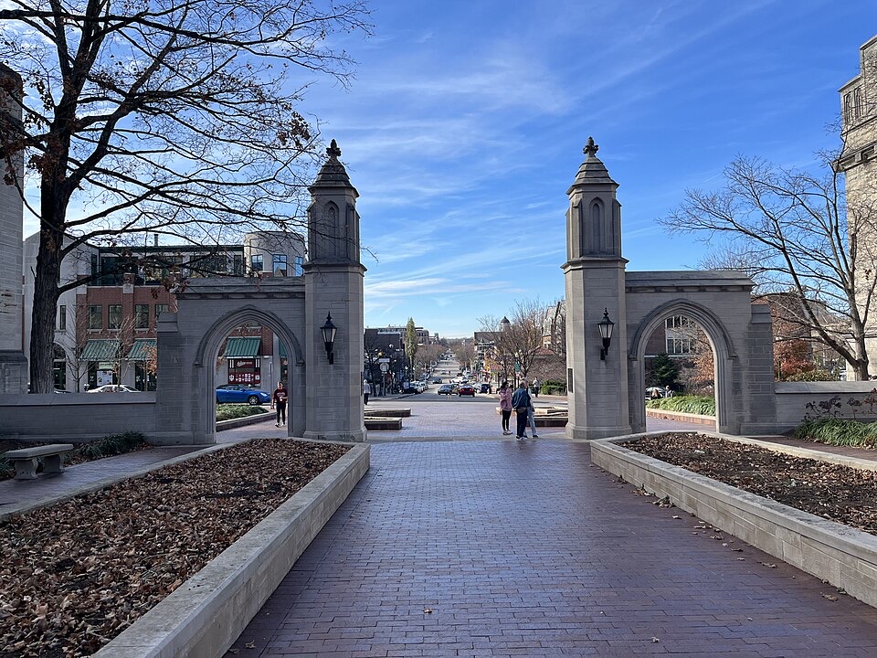 Stone archway entrance to a university campus with people walking through, surrounded by planters filled with fallen leaves. Buildings and a blue sky with wispy clouds are visible in the background.