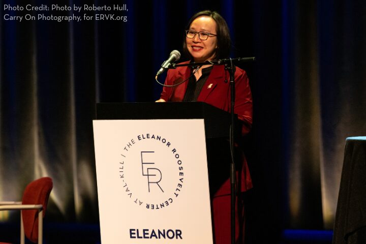 A woman in a maroon suit speaks at a podium with a microphone. The podium displays the Eleanor Roosevelt Center logo. The background is dark with vertical blue accents and there is a photo credit in the top left corner.