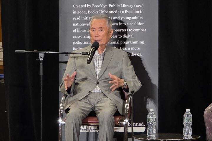 An older man in a gray suit sits and speaks into a microphone on stage. He gestures with his hands. Behind him is a banner with text about the Brooklyn Public Library’s Books Unbanned program. Two water bottles are visible nearby.