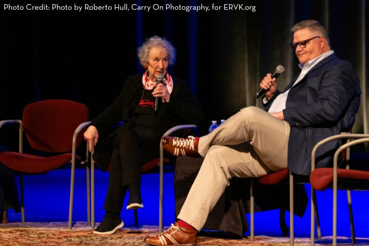 A woman and a man sit onstage in chairs, speaking into microphones during an event. The woman wears a black outfit and scarf; the man wears a suit with red sneakers. Stage lights cast a blue glow on the floor.